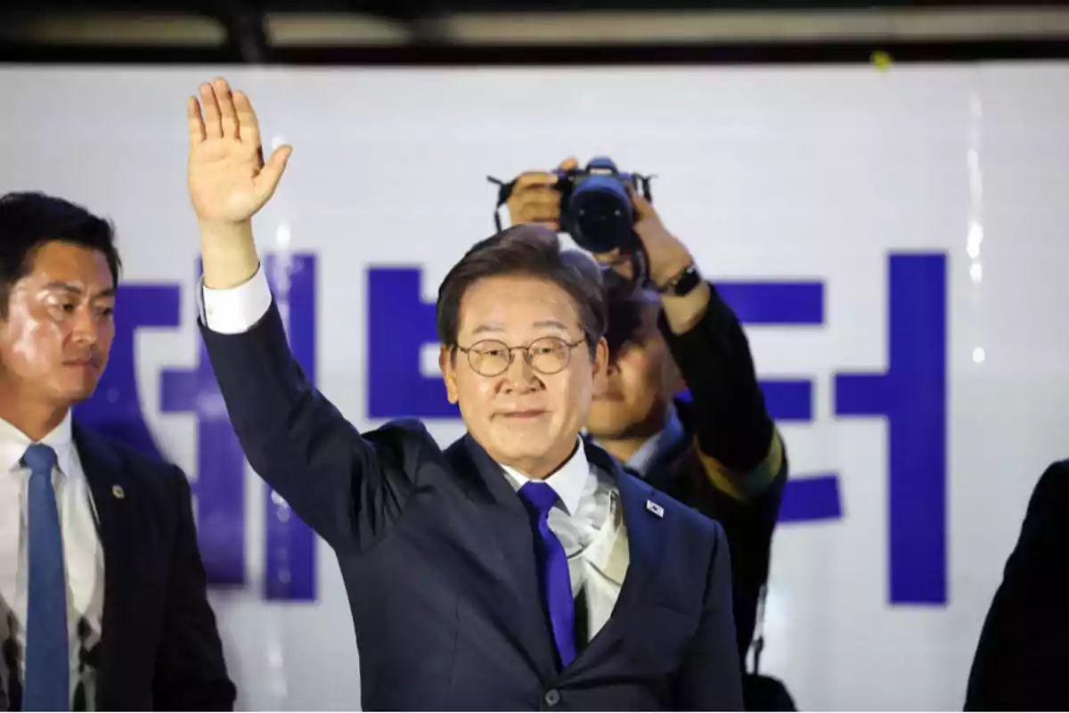 Hombre con gafas y traje azul saludando con la mano levantada mientras una persona toma fotos detrás de él