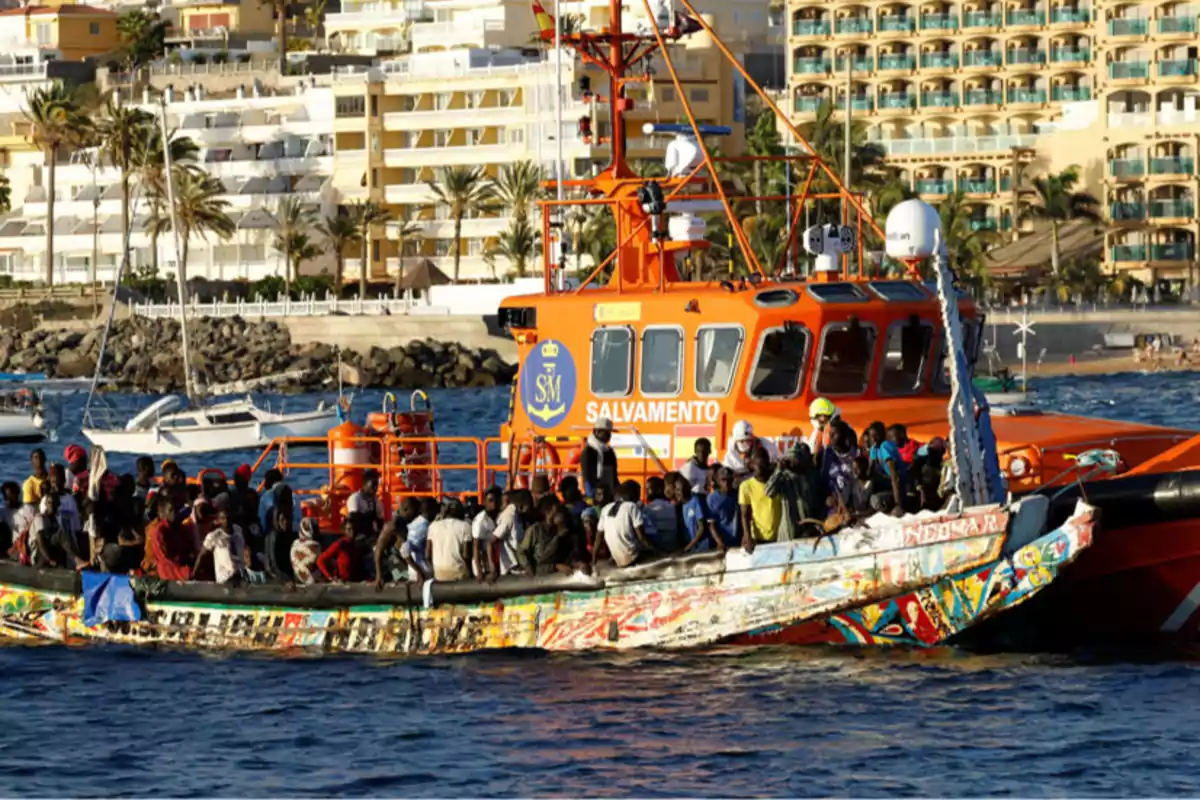 Un barco de rescate de Salvamento Marítimo ayuda a un grupo de personas en una embarcación sobrecargada cerca de la costa, con edificios y palmeras en el fondo.