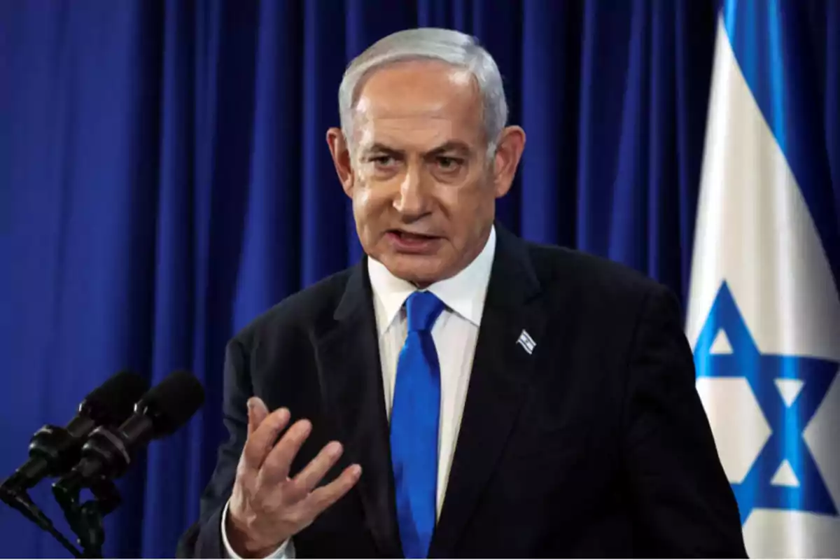 Un hombre de traje oscuro y corbata azul habla frente a micrófonos con una bandera de Israel al fondo.