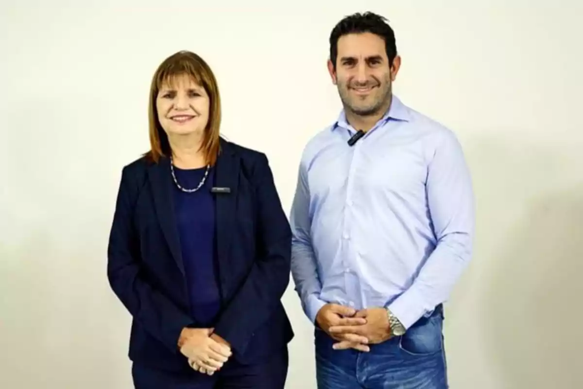Dos personas posando juntas, una mujer con traje oscuro y un hombre con camisa azul claro, ambos sonriendo frente a un fondo blanco.