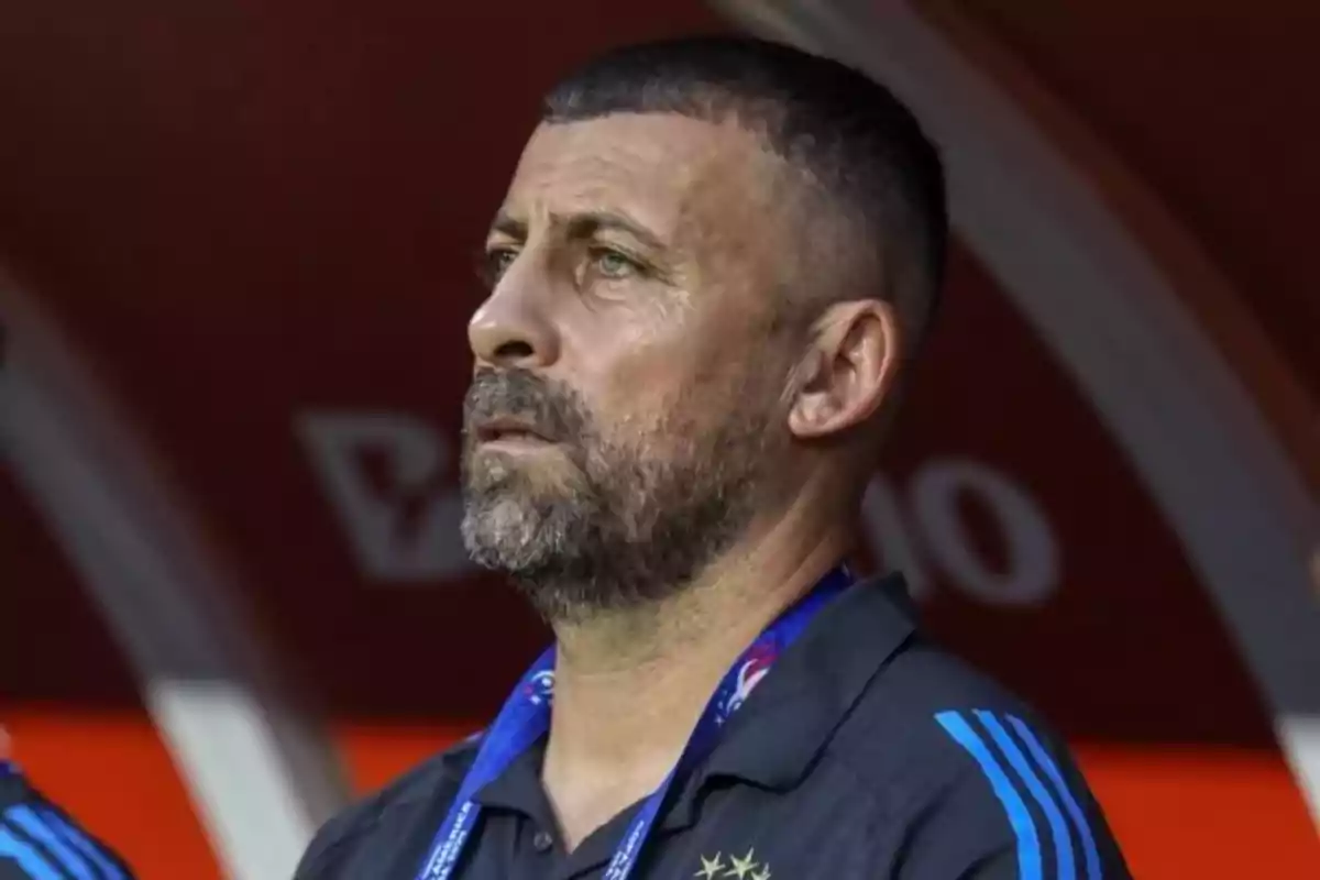 Short-haired, bearded man with a serious expression, wearing a dark sports jersey and a blue badge Short-haired, bearded man with a serious expression, wearing a dark sports jersey and a blue badge