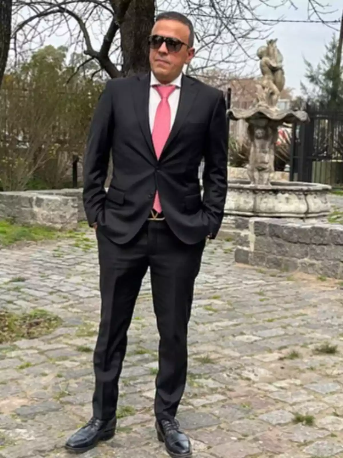 Man in a black suit and pink tie posing outdoors next to a stone fountain