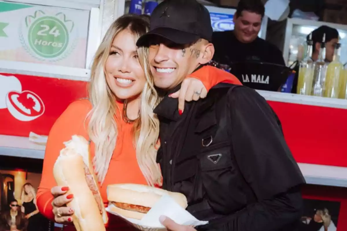 Una pareja sonriente sostiene sándwiches frente a un puesto de comida rápida. Una pareja sonriente sostiene sándwiches frente a un puesto de comida rápida.