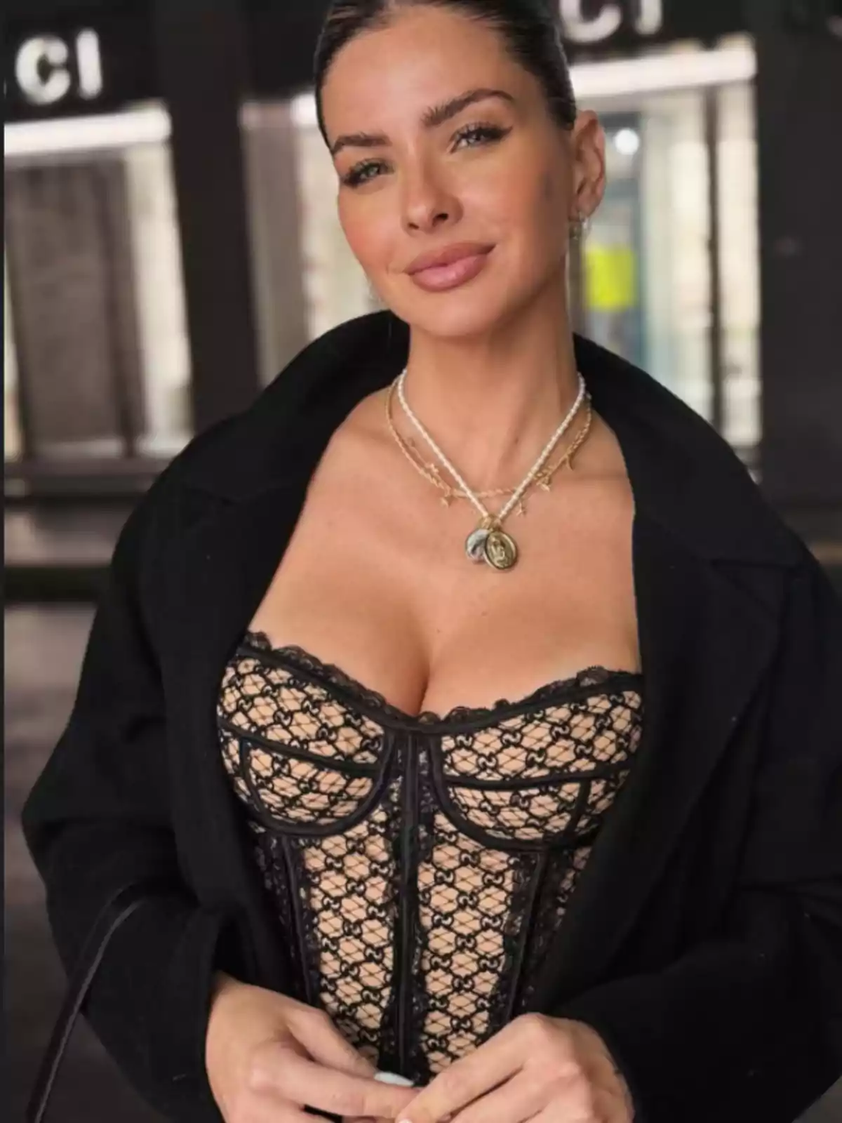 Mujer con el cabello recogido usando un corset de encaje negro y un abrigo oscuro, posando y sonriendo levemente frente a un fondo urbano desenfocado Mujer con el cabello recogido usando un corset de encaje negro y un abrigo oscuro, posando y sonriendo levemente frente a un fondo urbano desenfocado