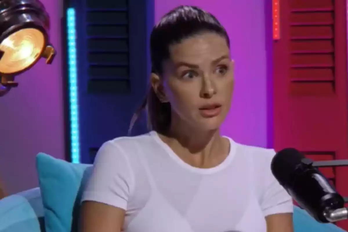 A person with a surprised expression is sitting in front of a microphone in a studio with colorful lights.