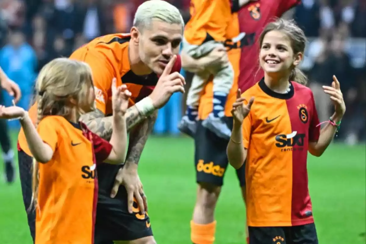 A player with platinum blonde hair interacts with two girls on the field, all wearing team jerseys in orange and red.
