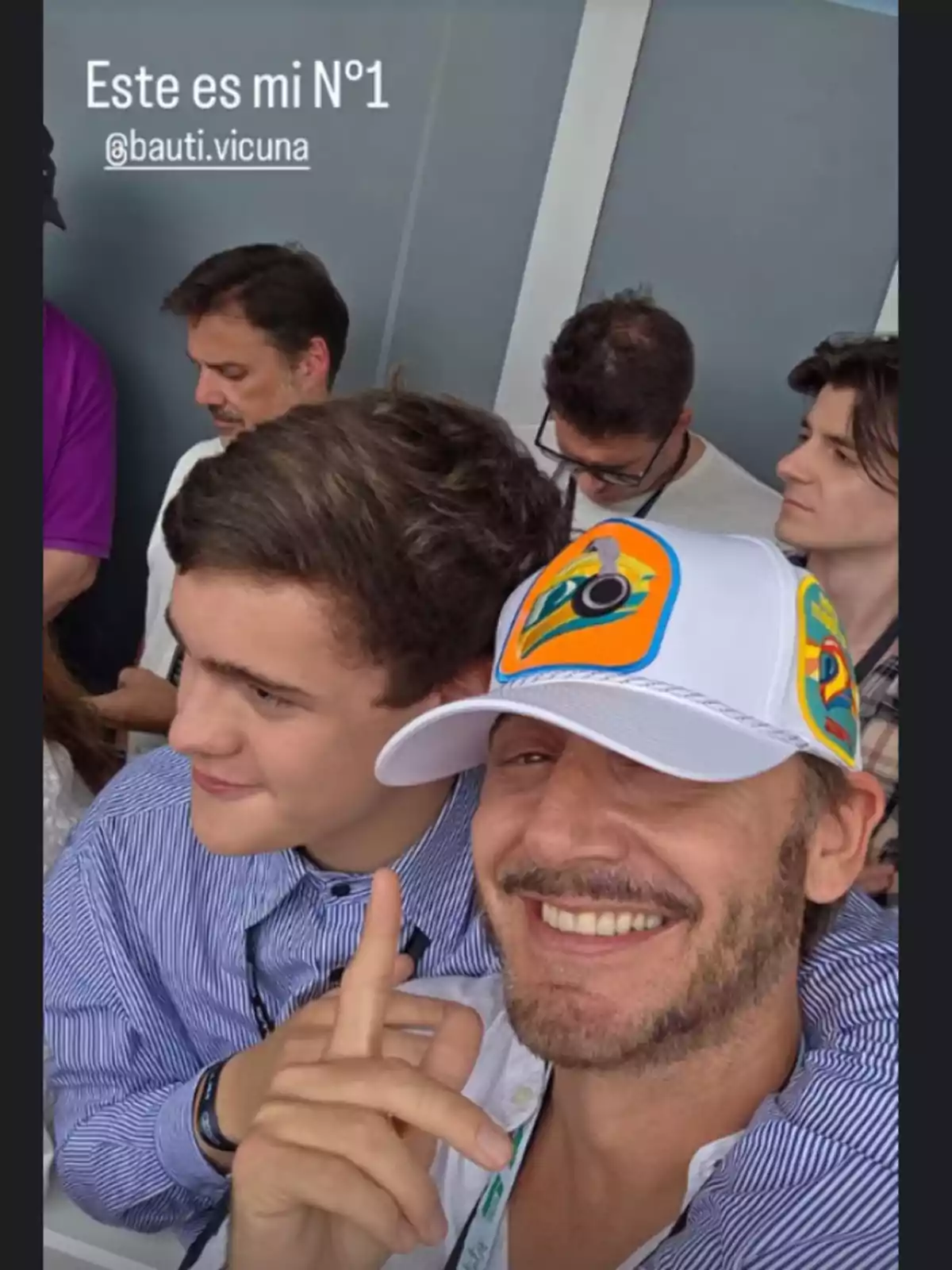 A group of people is gathered, with a man in the foreground smiling and pointing upwards, while wearing a white cap with a colorful design. A group of people is gathered, with a man in the foreground smiling and pointing upwards, while wearing a white cap with a colorful design.