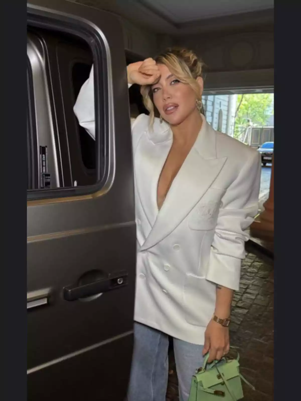 Una persona con un blazer blanco y jeans sostiene una puerta de auto mientras lleva un bolso verde. Una persona con un blazer blanco y jeans sostiene una puerta de auto mientras lleva un bolso verde.