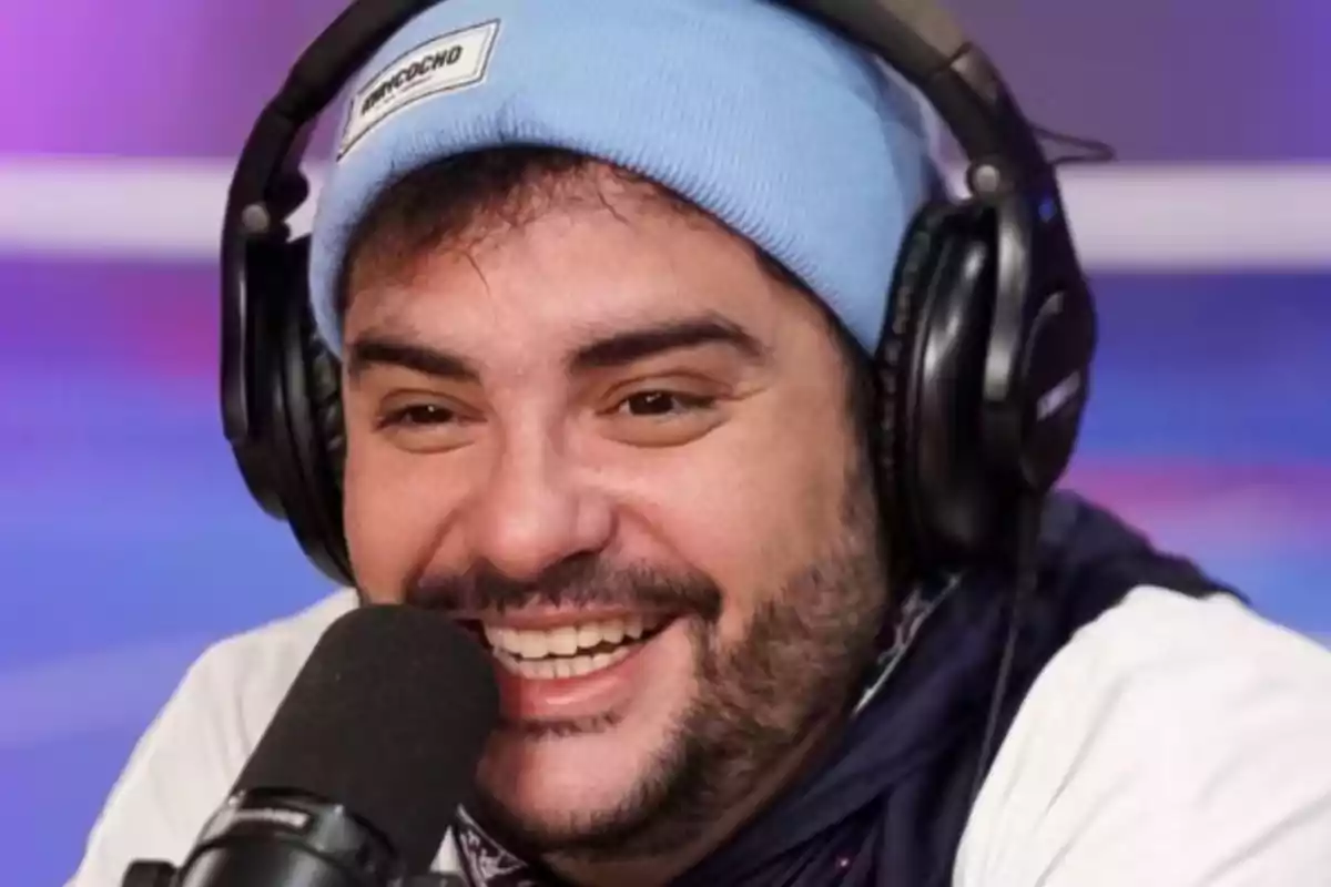 A smiling man with headphones and a blue beanie speaking in front of a microphone.