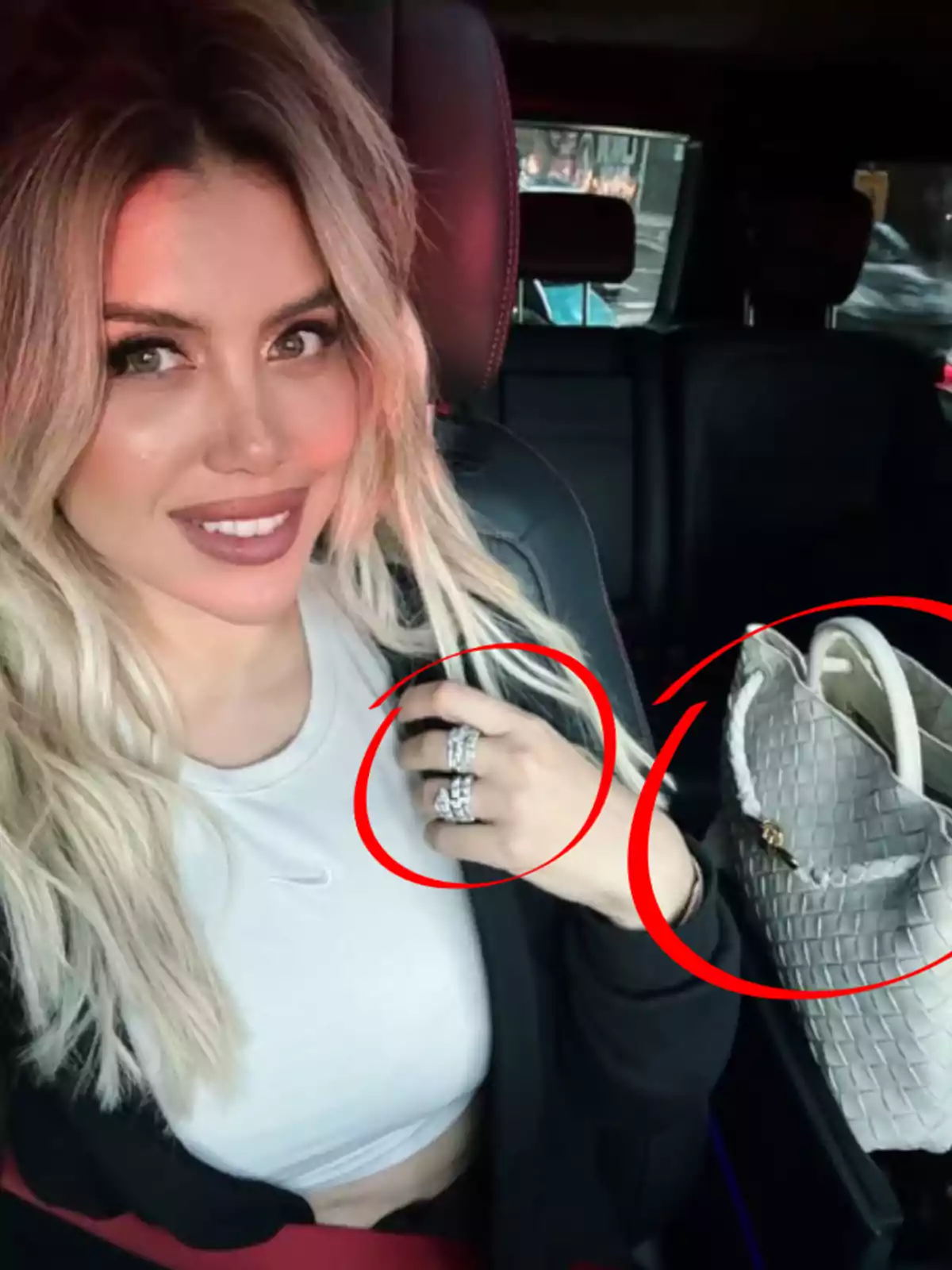 A person with blonde hair and makeup smiles while sitting in a vehicle, wearing a white top and a black jacket, with several shiny rings on their hand and a light-colored woven bag beside them. A person with blonde hair and makeup smiles while sitting in a vehicle, wearing a white top and a black jacket, with several shiny rings on their hand and a light-colored woven bag beside them.