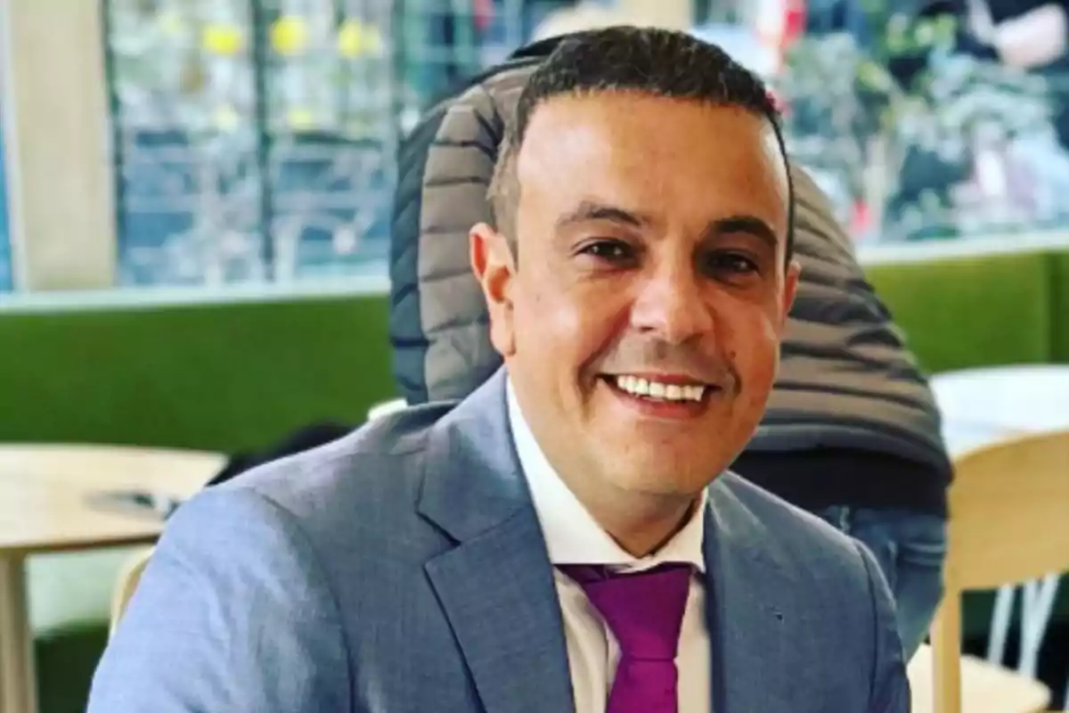 A smiling man in a gray suit and purple tie sitting in an indoor setting. A smiling man in a gray suit and purple tie sitting in an indoor setting.