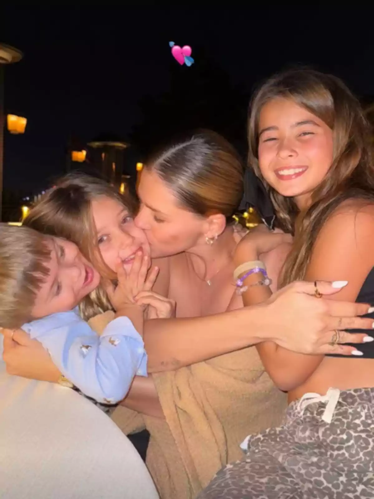 A woman hugs and affectionately kisses three children while they all smile on a night lit by warm lights. A woman hugs and affectionately kisses three children while they all smile on a night lit by warm lights.