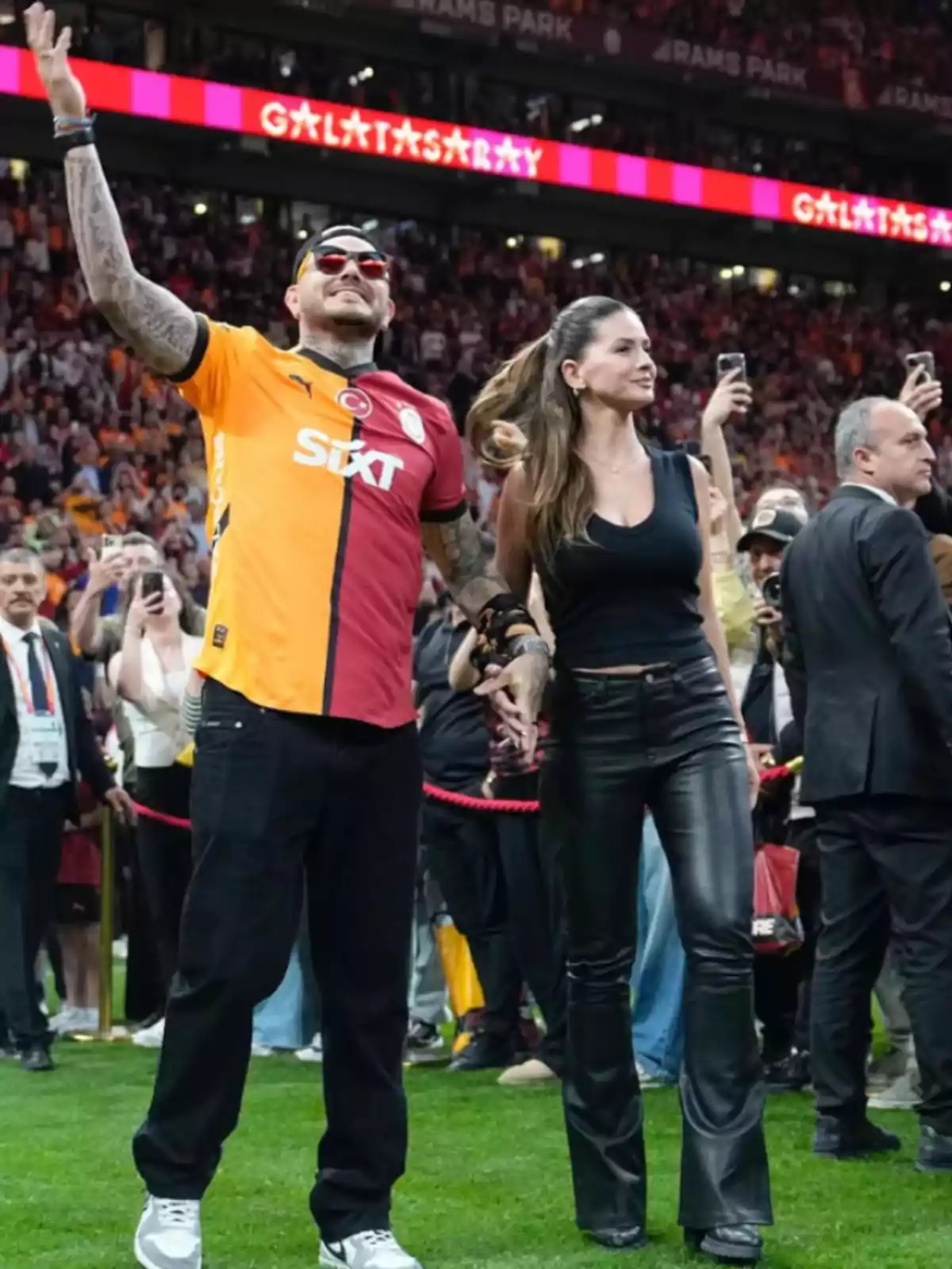 Un hombre con camiseta de fútbol del Galatasaray y una mujer de pantalón de cuero negro caminan tomados de la mano en un estadio lleno de gente Un hombre con camiseta de fútbol del Galatasaray y una mujer de pantalón de cuero negro caminan tomados de la mano en un estadio lleno de gente