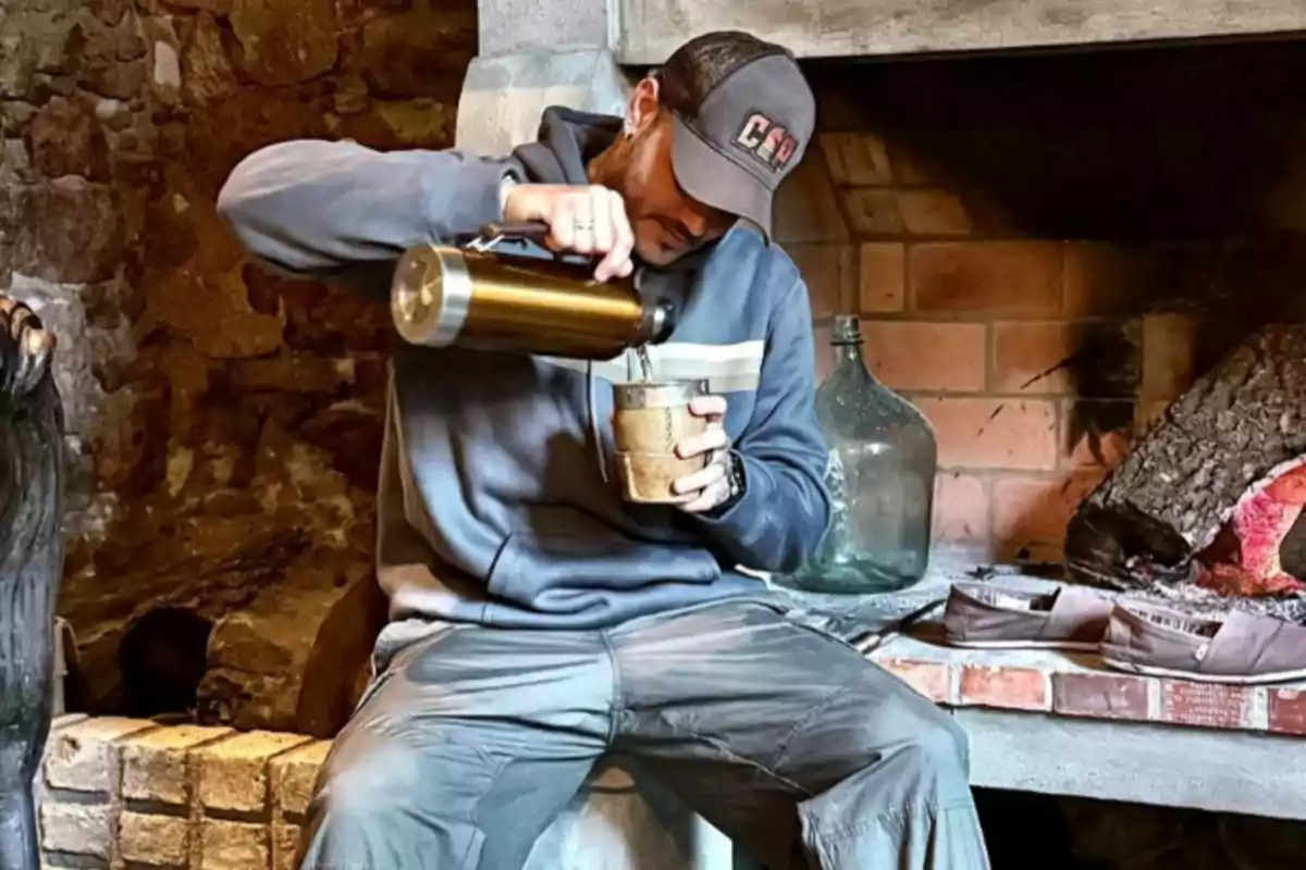 Hombre sentado junto a una chimenea sirviendo una bebida caliente en un vaso usando un termo mientras viste ropa casual y gorra Hombre sentado junto a una chimenea sirviendo una bebida caliente en un vaso usando un termo mientras viste ropa casual y gorra