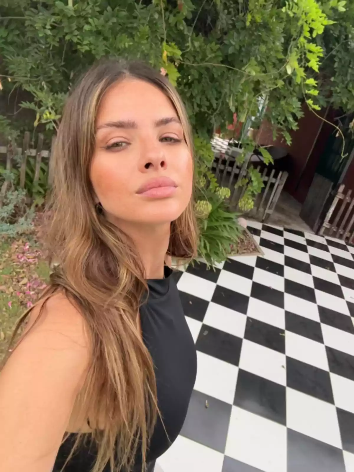 Mujer joven con cabello largo y suelto posando para una selfie en un patio con piso de cuadros blancos y negros y vegetación al fondo Mujer joven con cabello largo y suelto posando para una selfie en un patio con piso de cuadros blancos y negros y vegetación al fondo