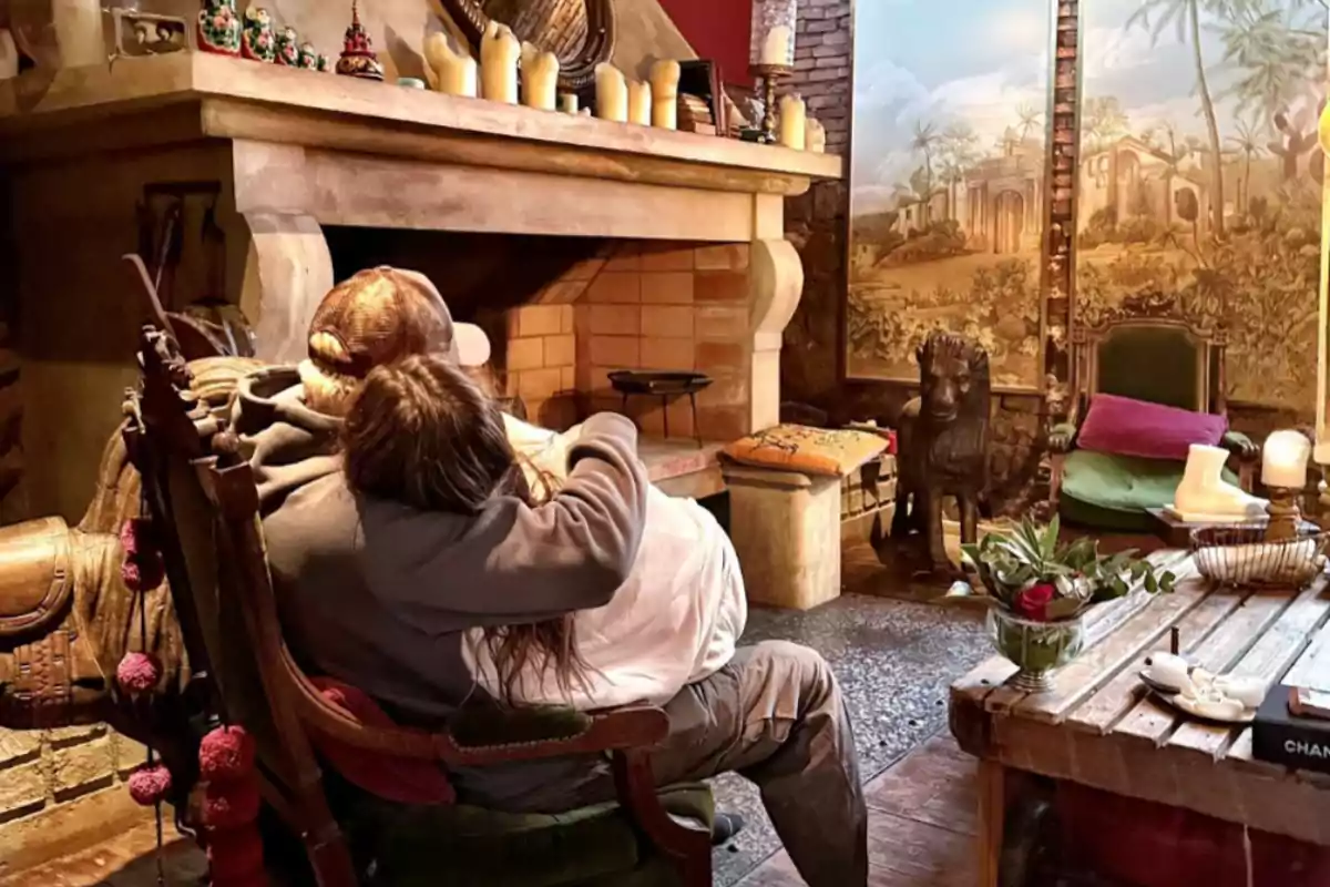 Pareja abrazada sentada frente a una chimenea en una sala acogedora decorada con velas, plantas y un mural de paisaje al fondo Pareja abrazada sentada frente a una chimenea en una sala acogedora decorada con velas, plantas y un mural de paisaje al fondo