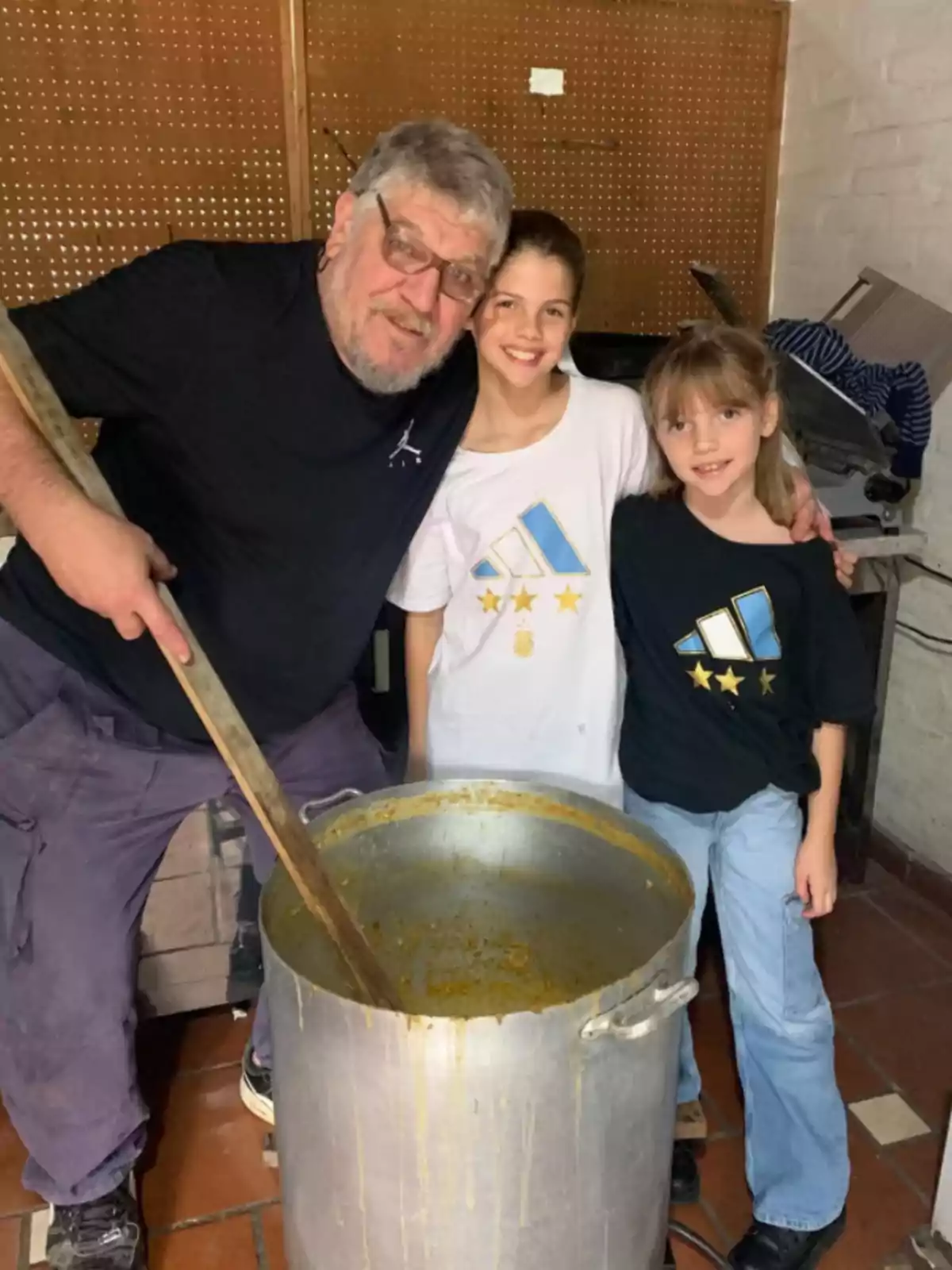 A smiling older man holds a large wooden spoon while cooking in a giant pot next to two girls who are also smiling and wearing t-jerseys with the Argentine national team crest. A smiling older man holds a large wooden spoon while cooking in a giant pot next to two girls who are also smiling and wearing t-jerseys with the Argentine national team crest.