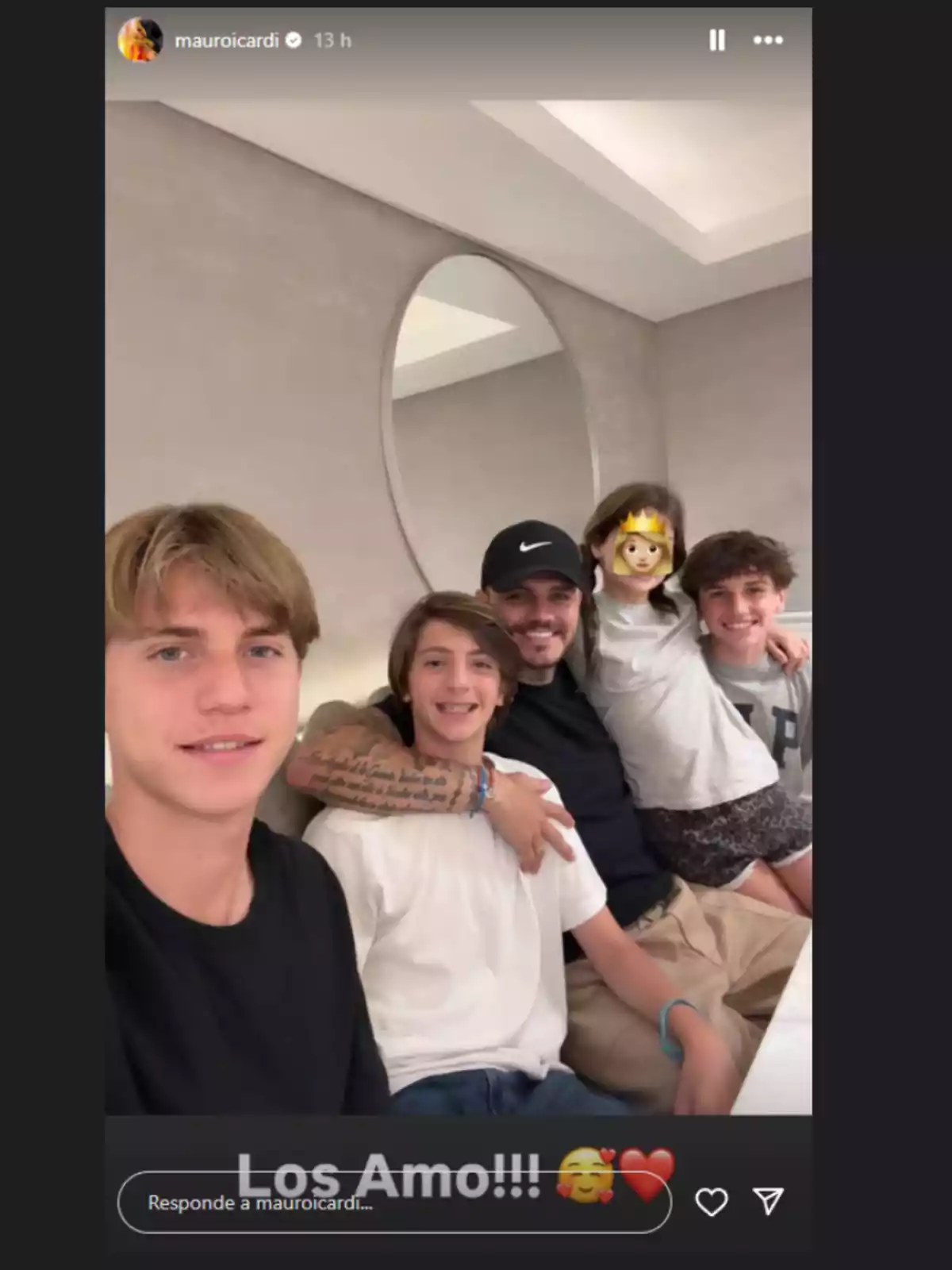 Cinco personas sentadas juntas en una mesa sonriendo, una niña tiene un emoji de princesa cubriendo su rostro y hay un espejo redondo en la pared de fondo Cinco personas sentadas juntas en una mesa sonriendo, una niña tiene un emoji de princesa cubriendo su rostro y hay un espejo redondo en la pared de fondo