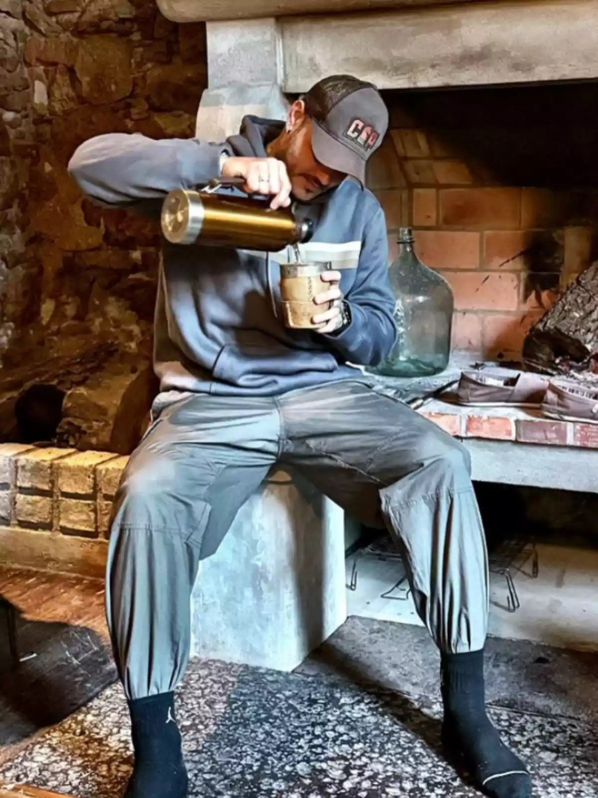 Hombre sentado frente a una chimenea sirviendo una bebida caliente en un vaso usando un termo mientras viste ropa cómoda y gorra Hombre sentado frente a una chimenea sirviendo una bebida caliente en un vaso usando un termo mientras viste ropa cómoda y gorra