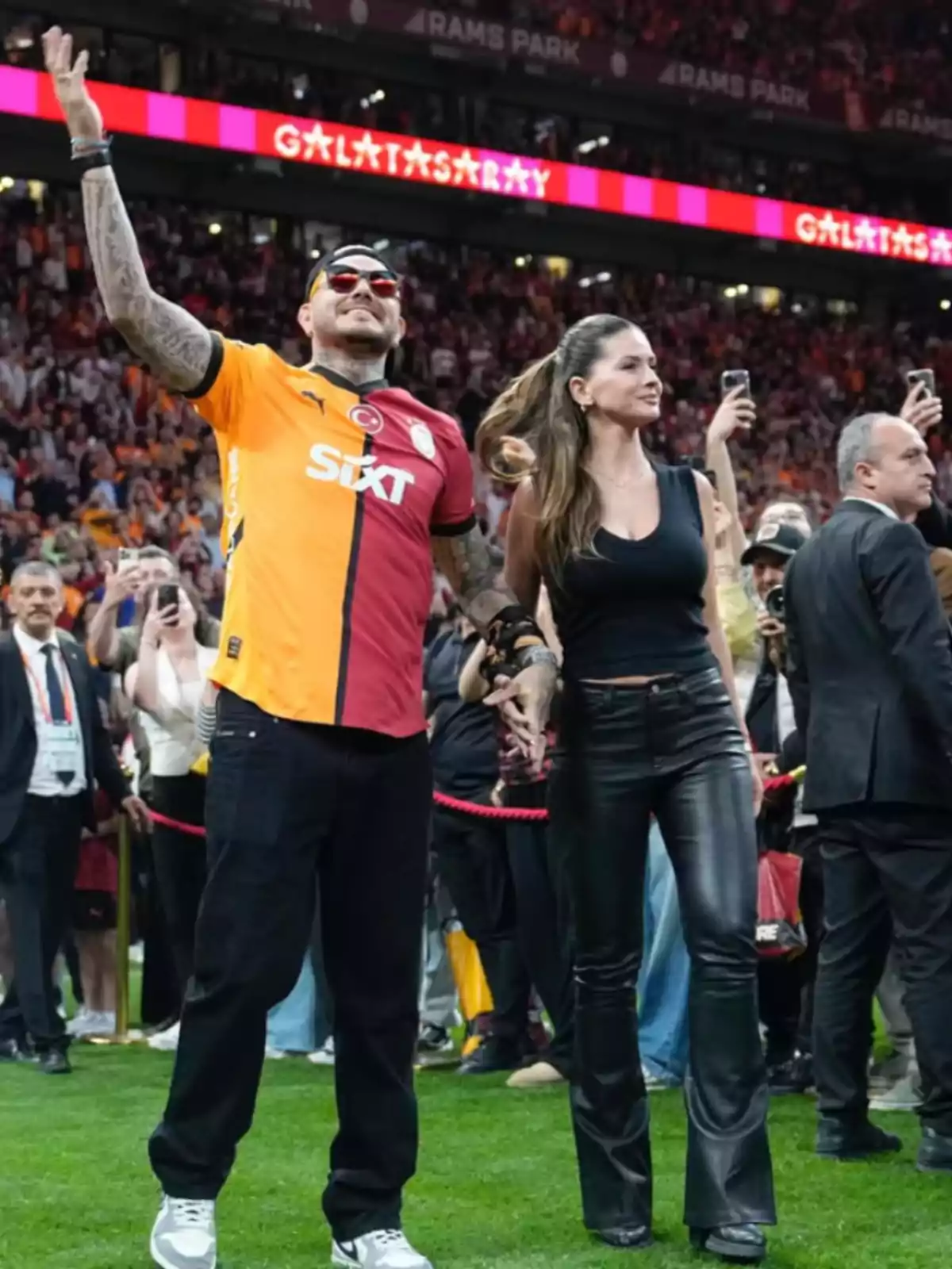 A man wearing a Galatasaray jersey and a woman in black leather pants walk hand in hand in a stadium full of fans. A man wearing a Galatasaray jersey and a woman in black leather pants walk hand in hand in a stadium full of fans.