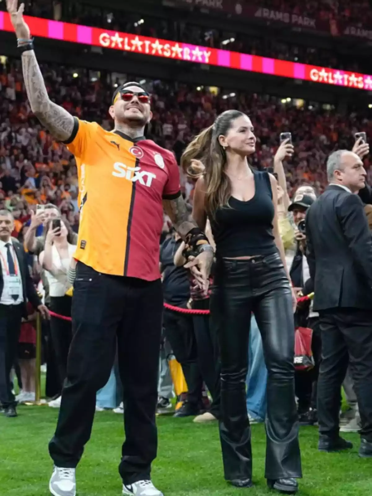 A man wearing a Galatasaray jersey and a woman in black leather pants walk hand in hand in a stadium full of fans. A man wearing a Galatasaray jersey and a woman in black leather pants walk hand in hand in a stadium full of fans.