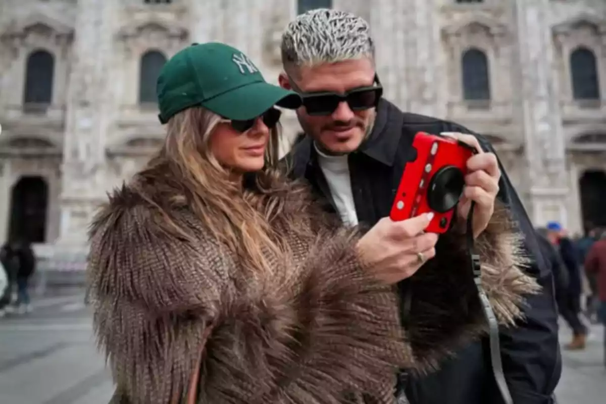 A couple with sunglasses watches a red camera while standing in front of a historic building.