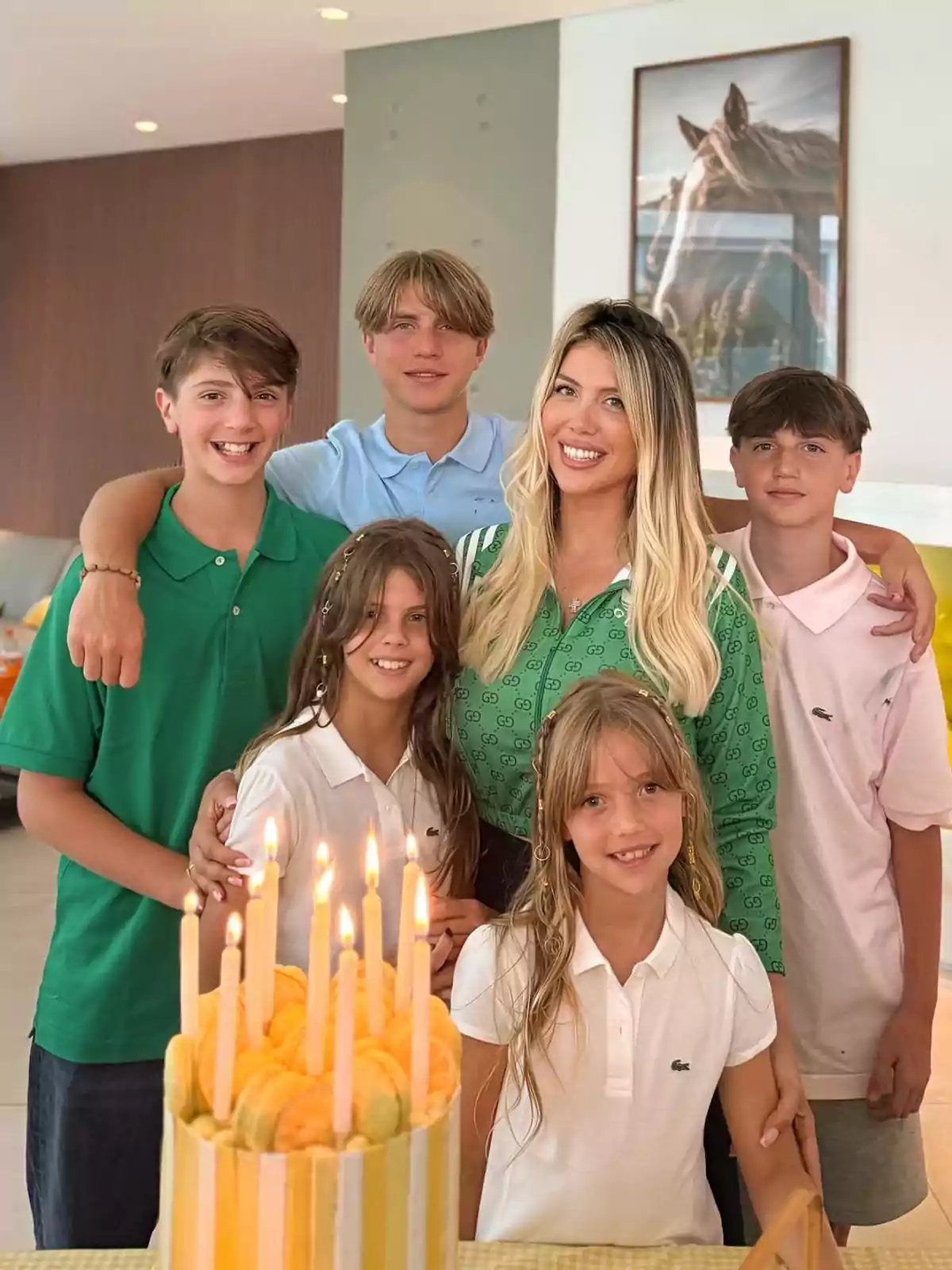 Grupo de cinco niños y una mujer sonriente posando juntos detrás de un pastel de cumpleaños con velas encendidas en un ambiente interior decorado con un cuadro de un caballo al fondo Grupo de cinco niños y una mujer sonriente posando juntos detrás de un pastel de cumpleaños con velas encendidas en un ambiente interior decorado con un cuadro de un caballo al fondo