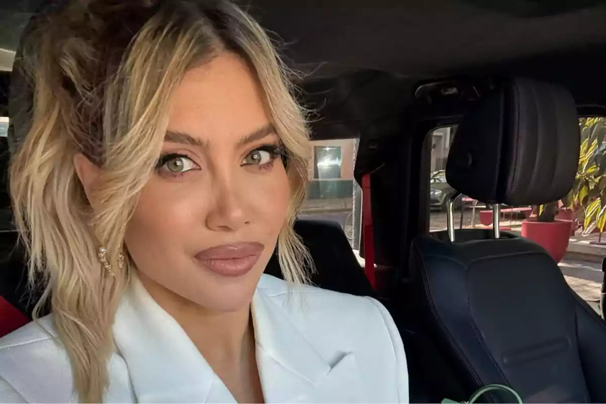 Blonde woman with makeup and a white jacket sitting inside a car looking at the camera