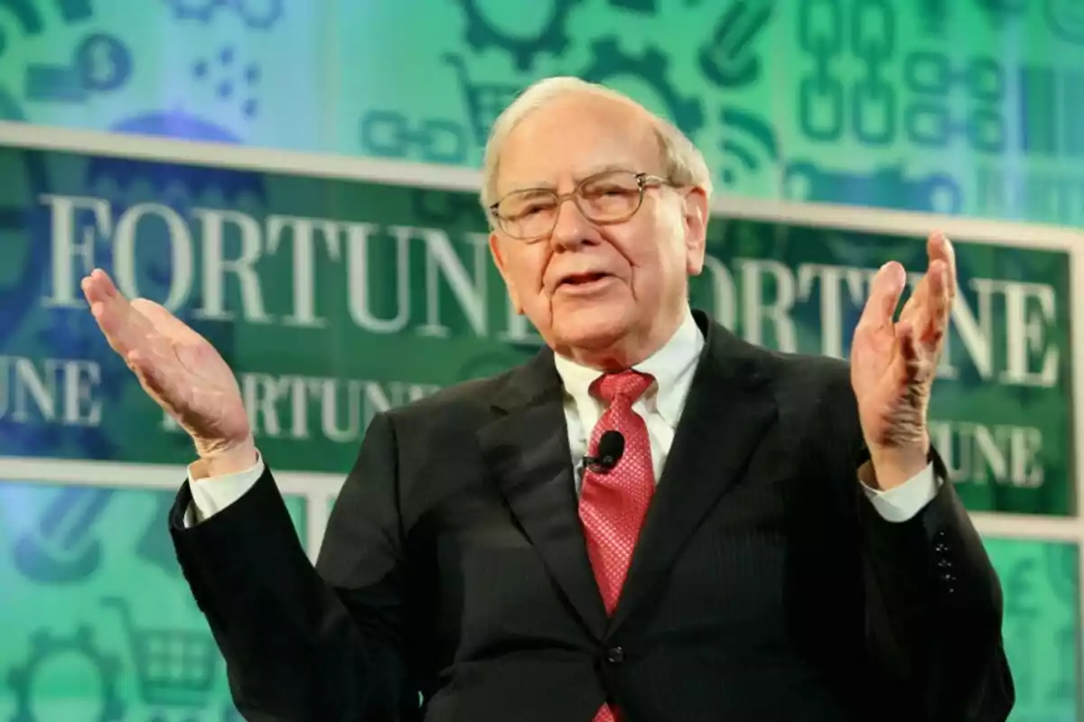 An older man with glasses and a dark suit gestures while speaking at an event with a green background that has the word An older man with glasses and a dark suit gestures while speaking at an event with a green background that has the word