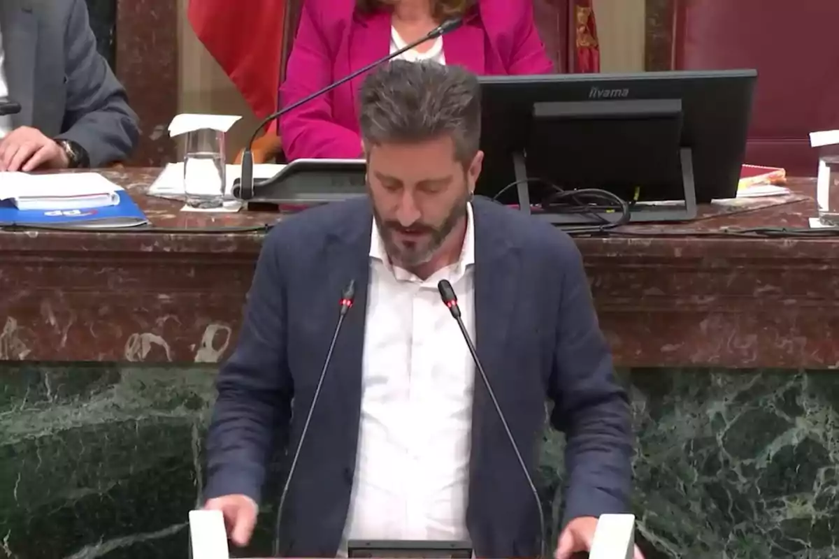 Man standing and speaking at a podium in a government room with microphones and people seated in the background