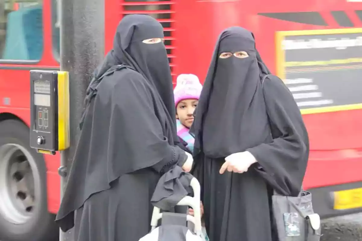 Two women wearing black niqabs and a girl with a pink hat are on a street next to a pedestrian traffic light and a red bus. Two women wearing black niqabs and a girl with a pink hat are on a street next to a pedestrian traffic light and a red bus.