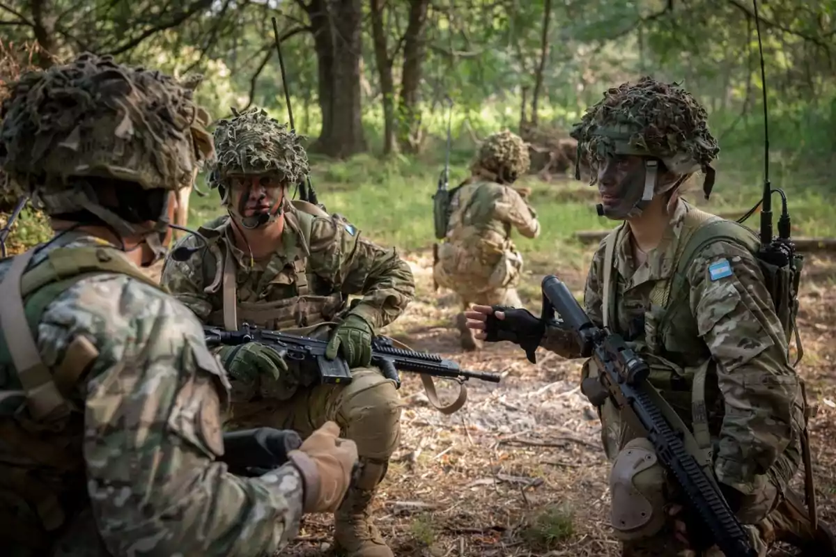 Un grupo de soldados con uniformes de camuflaje y cascos se encuentra en un bosque, algunos están agachados y sostienen rifles mientras conversan y parecen estar en una operación táctica. Un grupo de soldados con uniformes de camuflaje y cascos se encuentra en un bosque, algunos están agachados y sostienen rifles mientras conversan y parecen estar en una operación táctica.