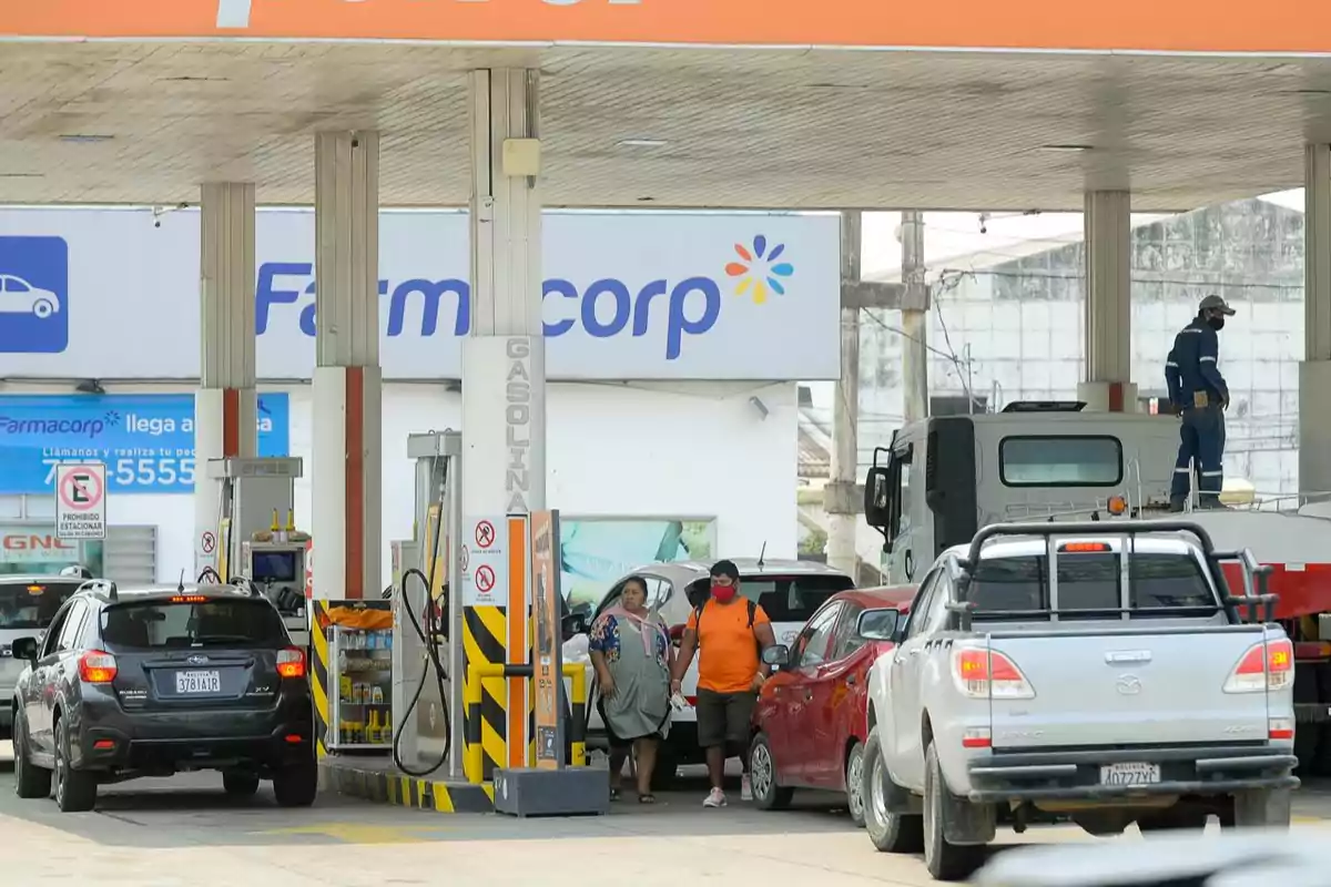 Una estación de servicio con varios vehículos en fila para cargar combustible y personas caminando cerca de las bombas.