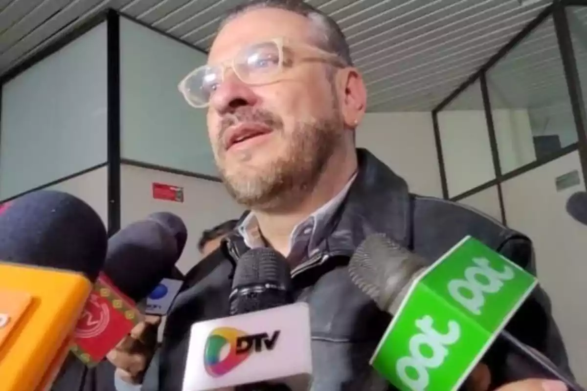 Un hombre con gafas y barba está rodeado de micrófonos de diferentes medios de comunicación en un entorno interior.