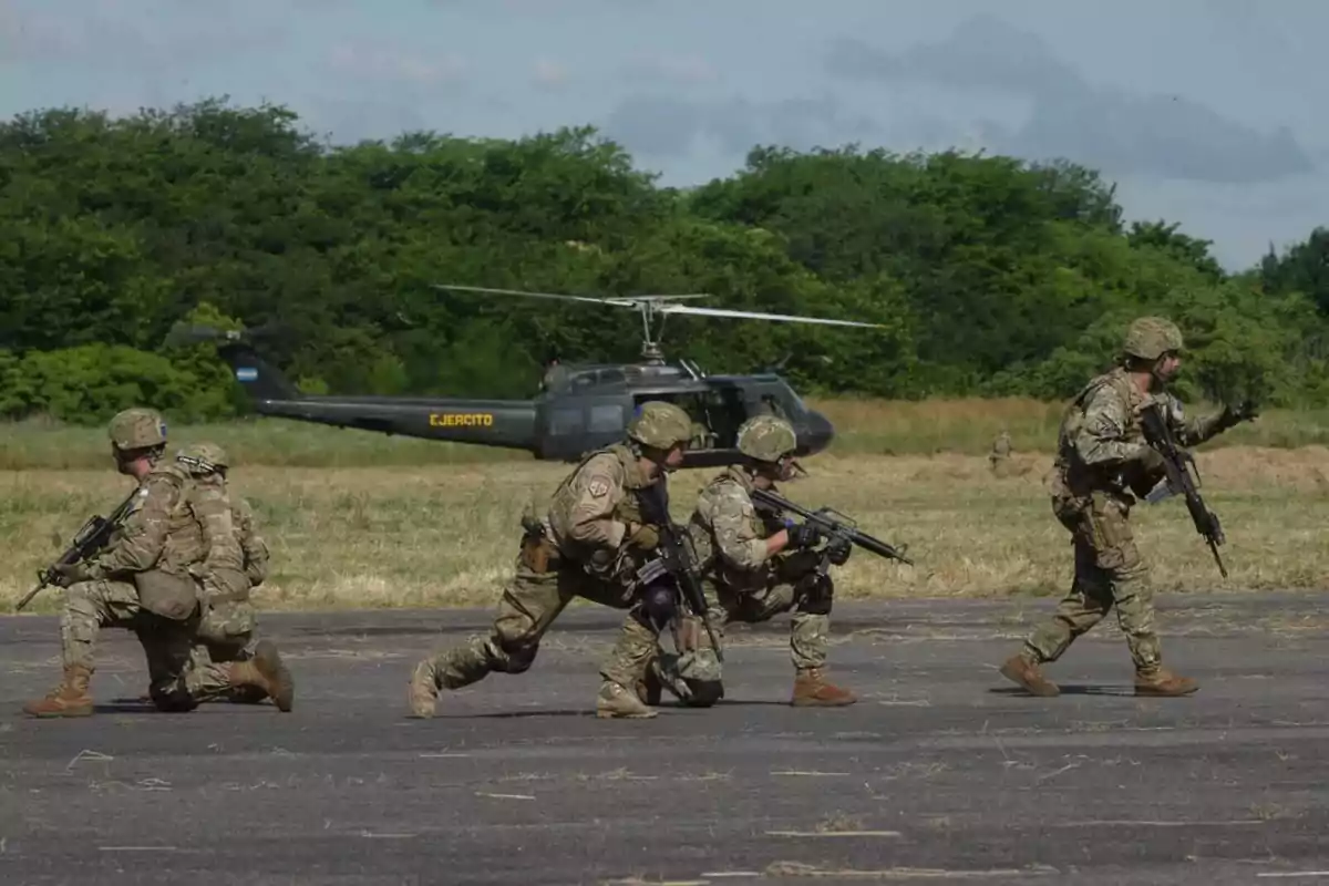 Soldados en uniforme militar con armas avanzan en formación frente a un helicóptero del ejército en un campo abierto.