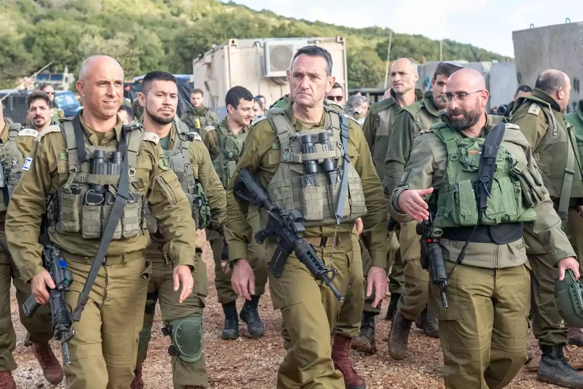 Un grupo de soldados con uniformes militares camina juntos en un entorno al aire libre, algunos llevan rifles y chalecos tácticos.