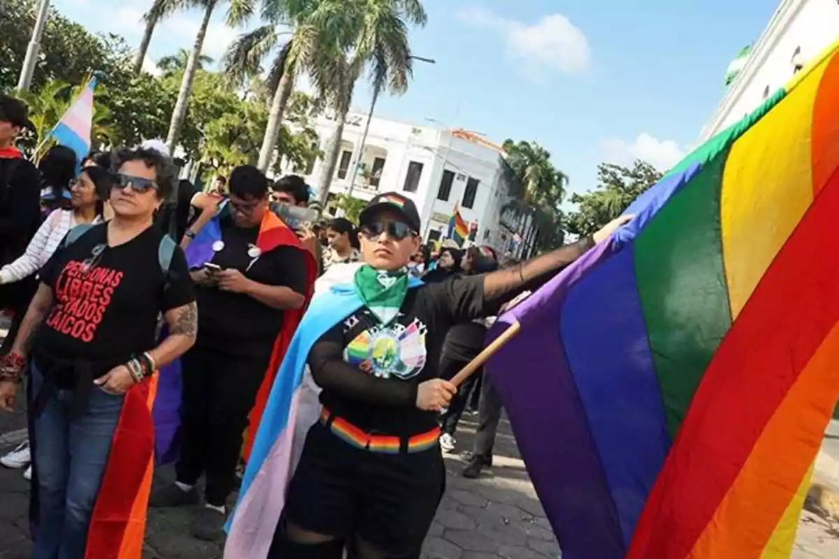 Personas marchando en una manifestación del orgullo LGBT con banderas de colores y ropa alusiva a la diversidad sexual en una calle rodeada de palmeras y edificios blancos