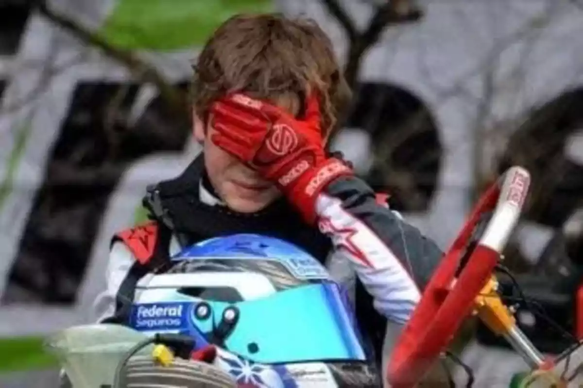 Un joven piloto de karting con guantes rojos se cubre la cara con una mano mientras sostiene su casco azul.