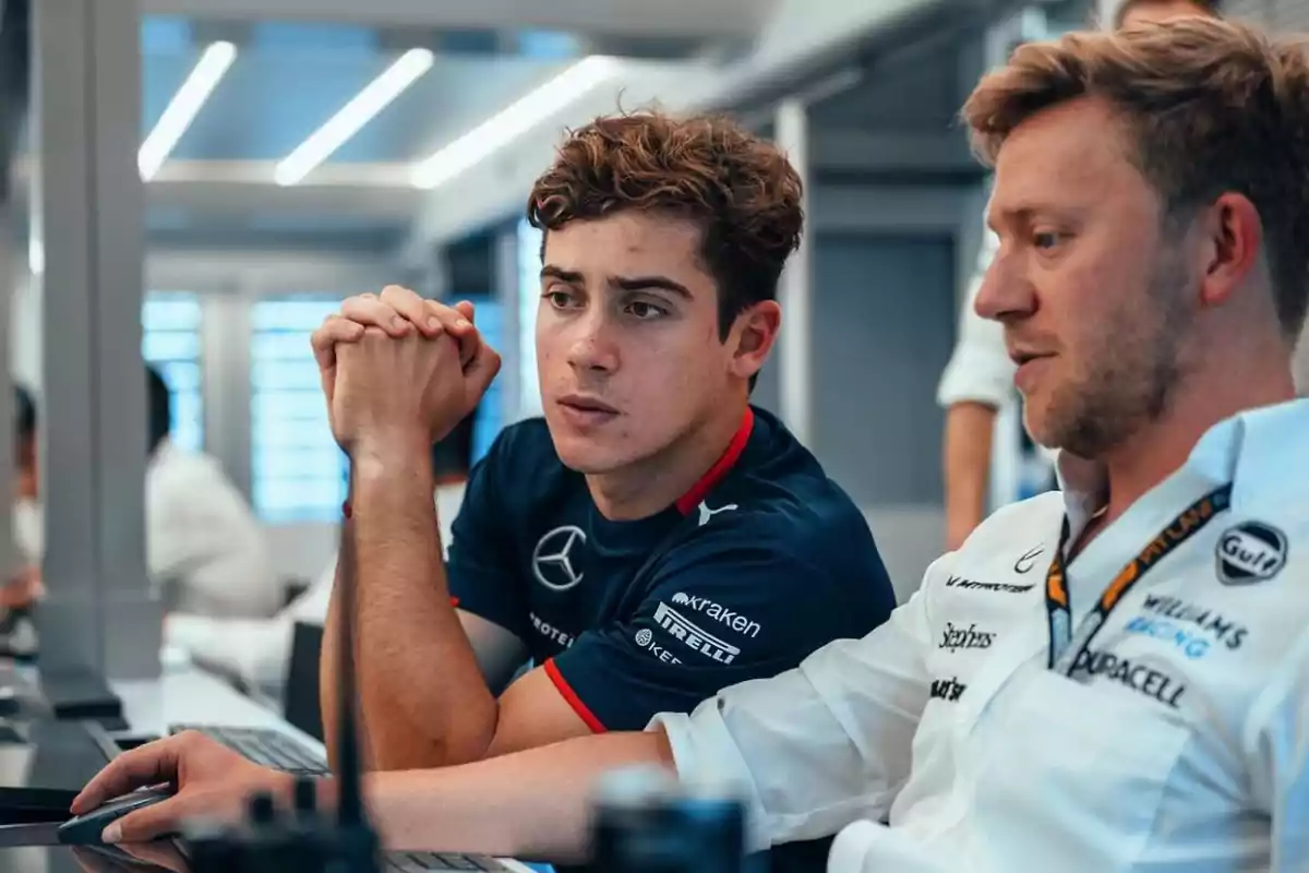 Dos hombres trabajando juntos en una oficina, uno de ellos lleva una camiseta de Mercedes y el otro una camisa blanca con logotipos de patrocinadores.