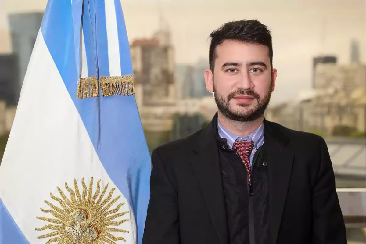 Un hombre de traje y corbata posa junto a una bandera de Argentina con un fondo urbano desenfocado.