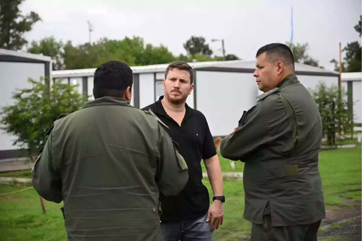 Un hombre de pie hablando con dos oficiales en uniforme verde en un entorno al aire libre con edificios modulares al fondo.