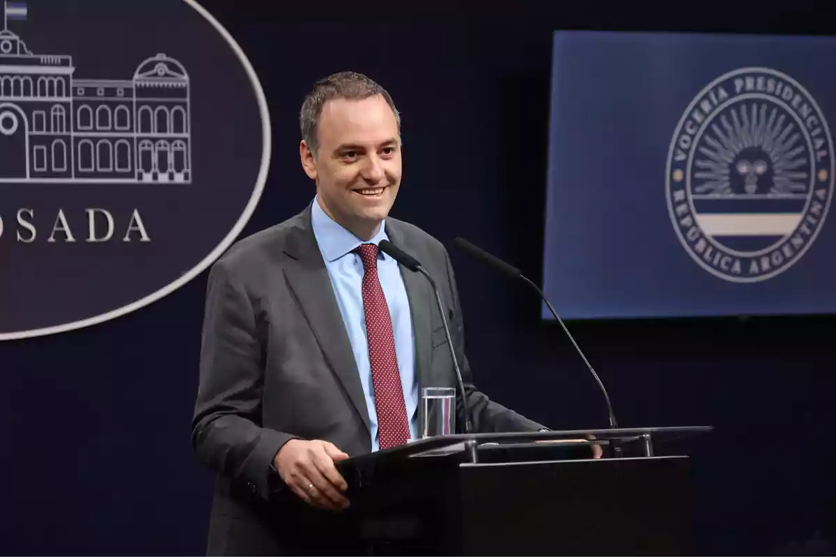 Un hombre de traje y corbata roja está hablando en un podio con micrófonos, con un fondo que muestra el logo de la Casa Rosada y la Vocería Presidencial de la República Argentina.