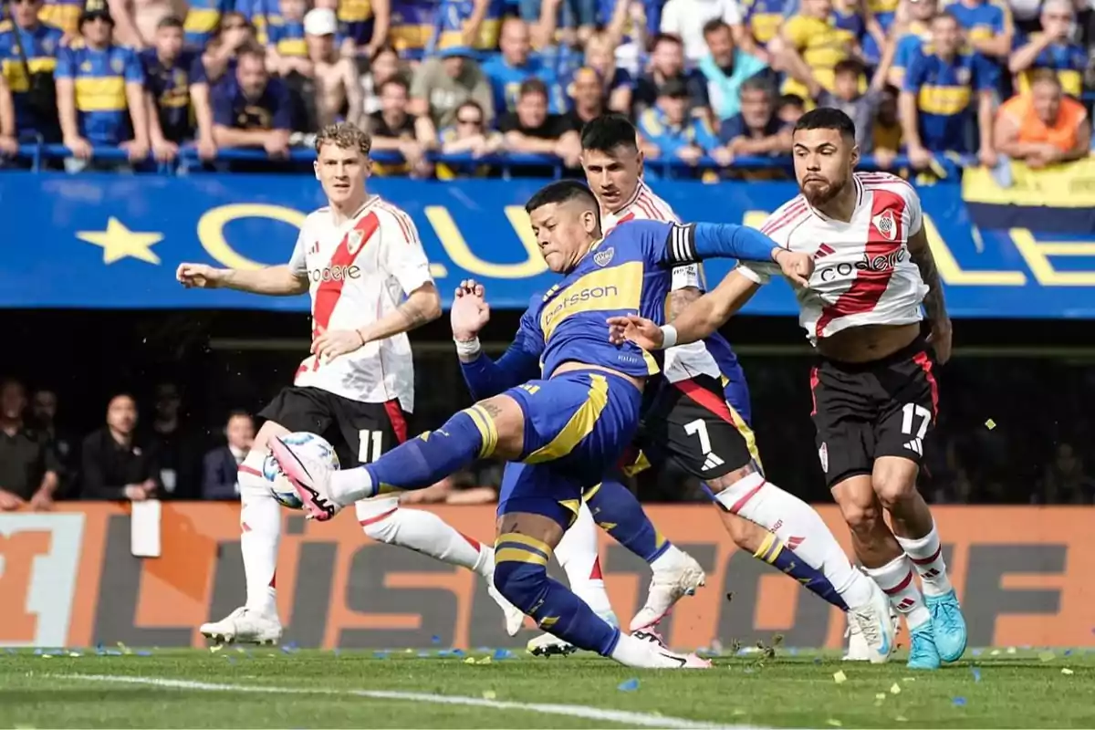 Jugadores de Boca Juniors y River Plate disputando un balón durante un partido de fútbol con aficionados en las gradas.