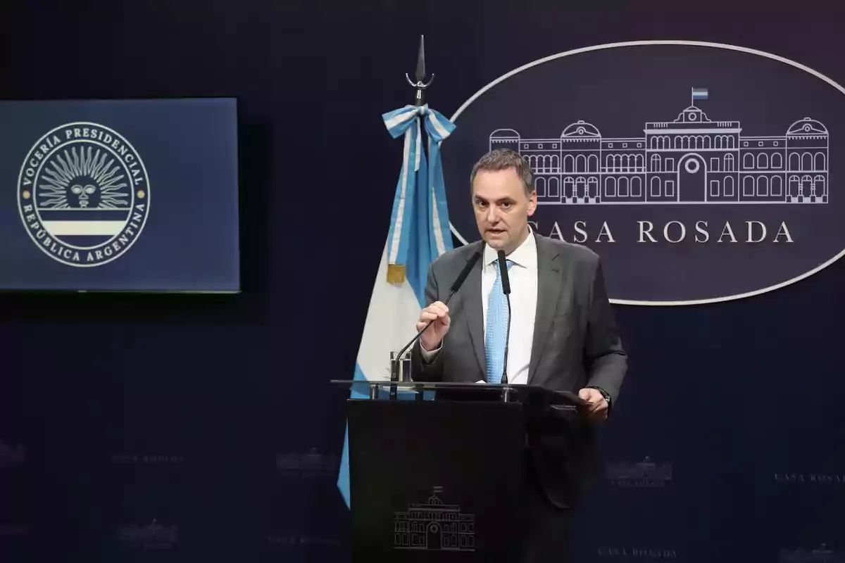 Un hombre en traje habla en un podio con el logo de la Casa Rosada y la bandera de Argentina detrás.