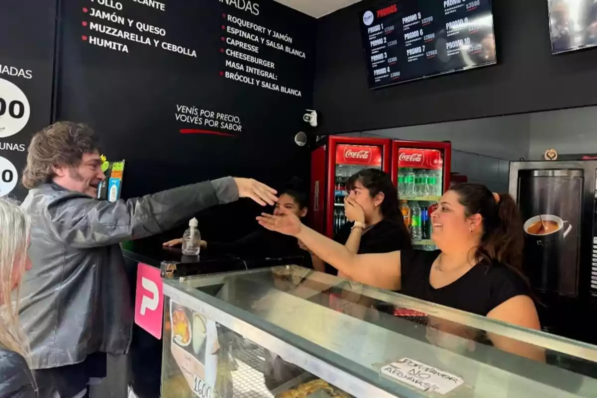 Un hombre con chaqueta de cuero interactúa con tres mujeres detrás de un mostrador en un local de comida rápida, mientras una de ellas sonríe y otra se cubre la boca sorprendida.