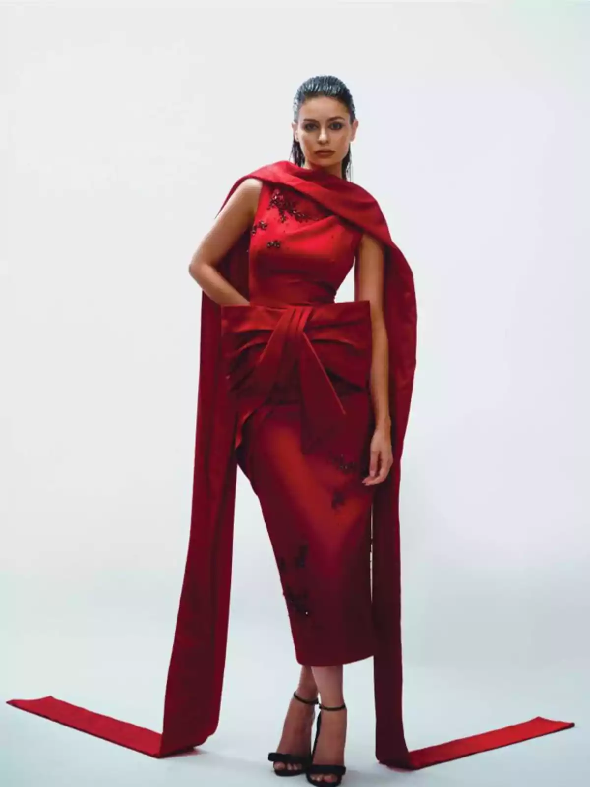 Mujer con vestido rojo elegante y detalles negros, con un diseño de lazo grande y tela larga, posando en un fondo blanco.