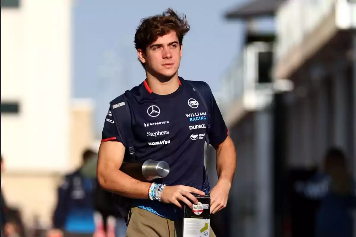 Un joven con camiseta de Williams Racing camina al aire libre sosteniendo una botella y una caja.