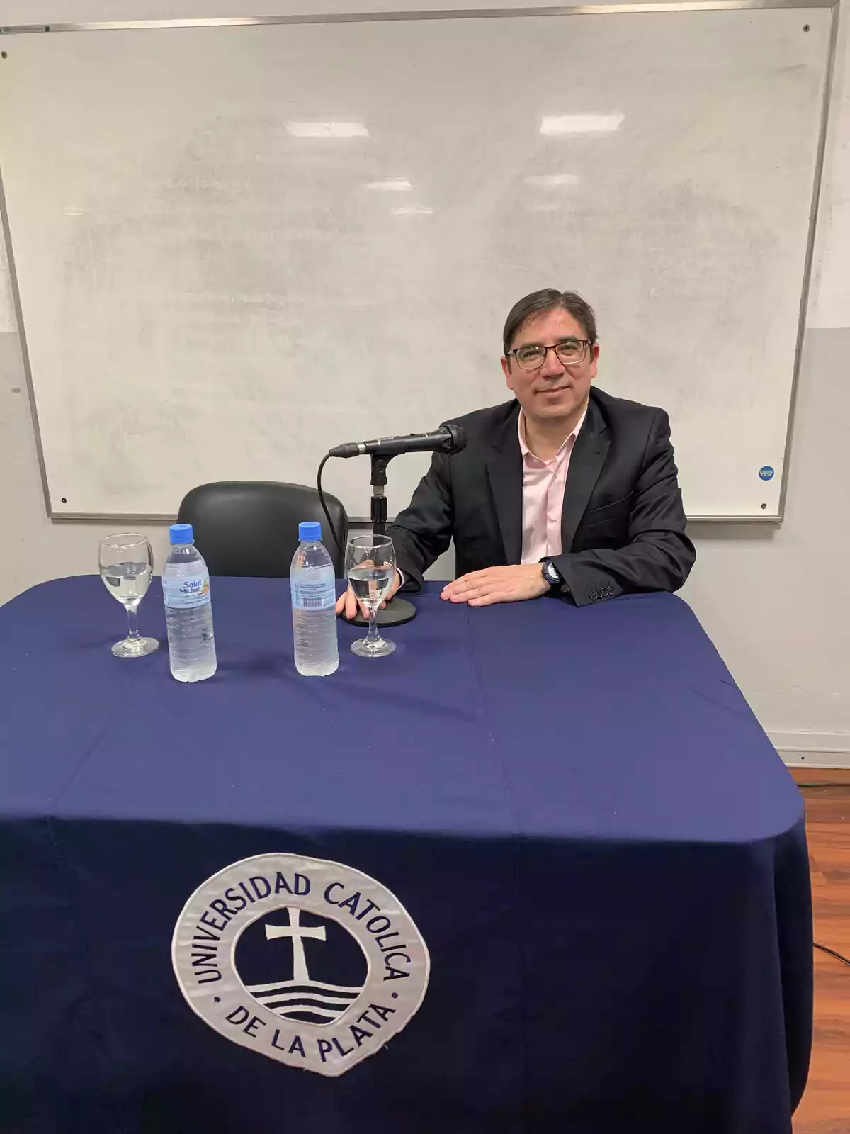 Hombre sentado en una mesa con un mantel de la Universidad Católica de La Plata, frente a un micrófono y dos botellas de agua.