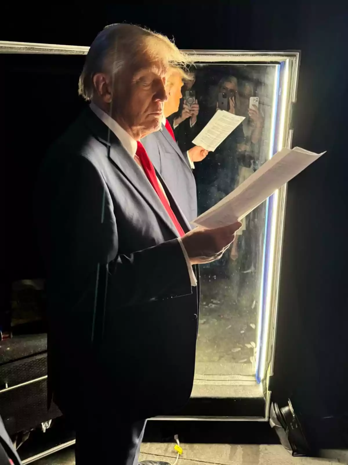 Un hombre de traje oscuro y corbata roja sostiene un documento mientras se refleja en un espejo iluminado.