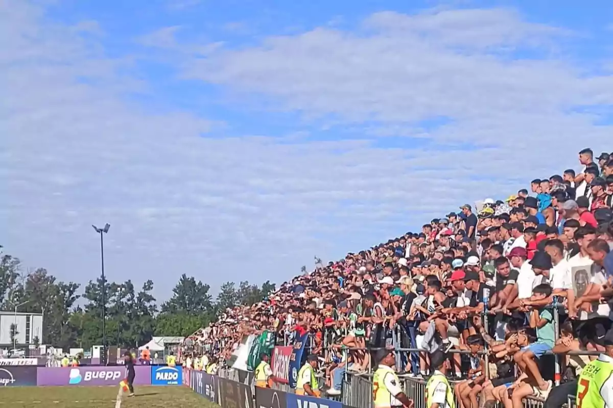 Una multitud de personas llenan las gradas de un estadio al aire libre bajo un cielo azul con nubes dispersas.